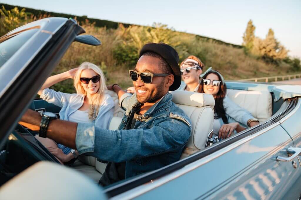 Group of cheerful young friends driving car and smiling under a summer season