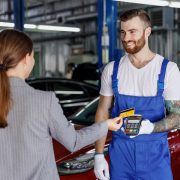 Young happy cheerful professional car mechanic man in blue overalls gloves hold payment terminal fow paying with credit card by female owner driver woman work in vehicle repair shop workshop indoors.