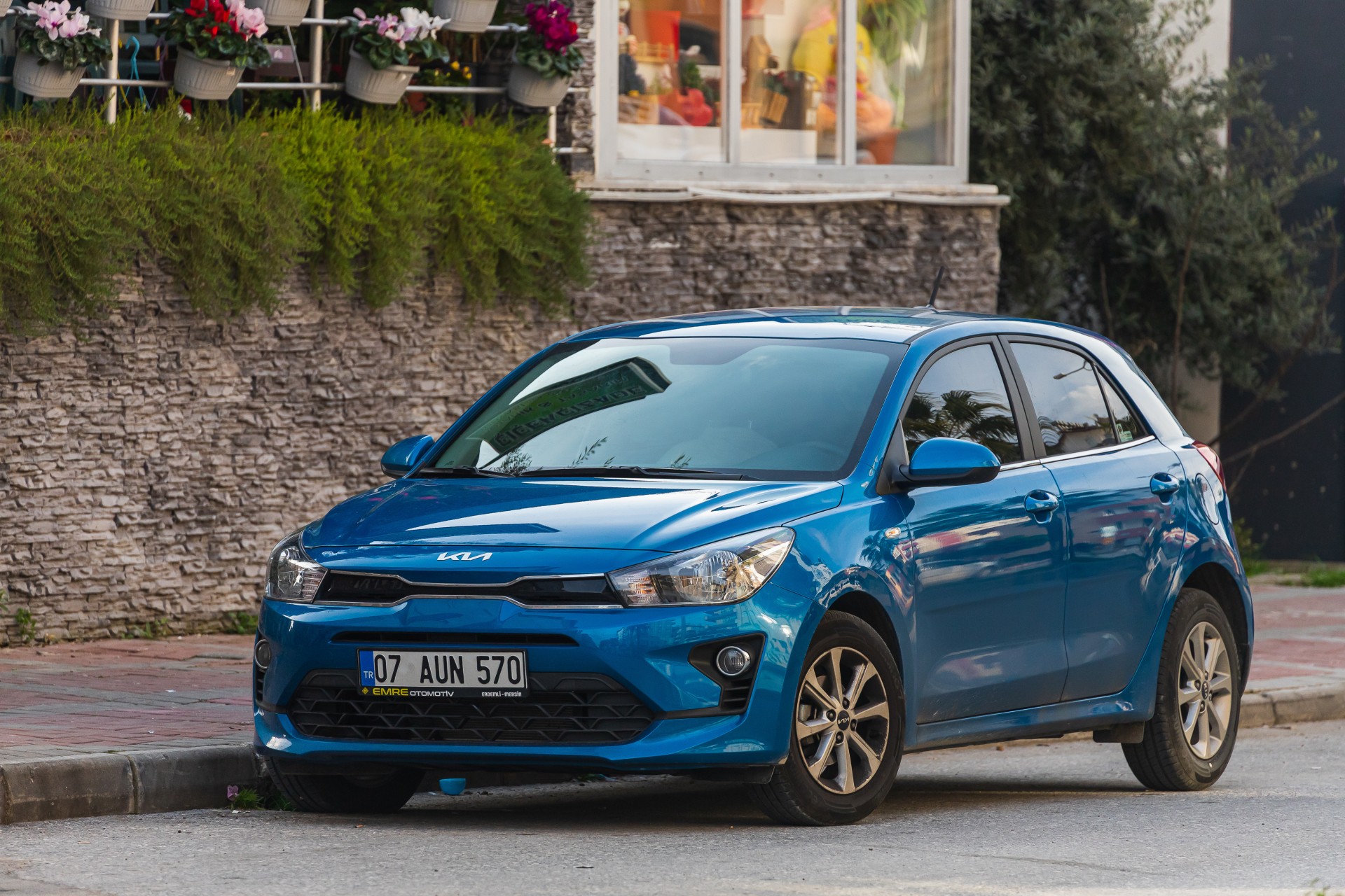 blue Kia Rio is parked on the street on a warm day against the backdrop of a garden