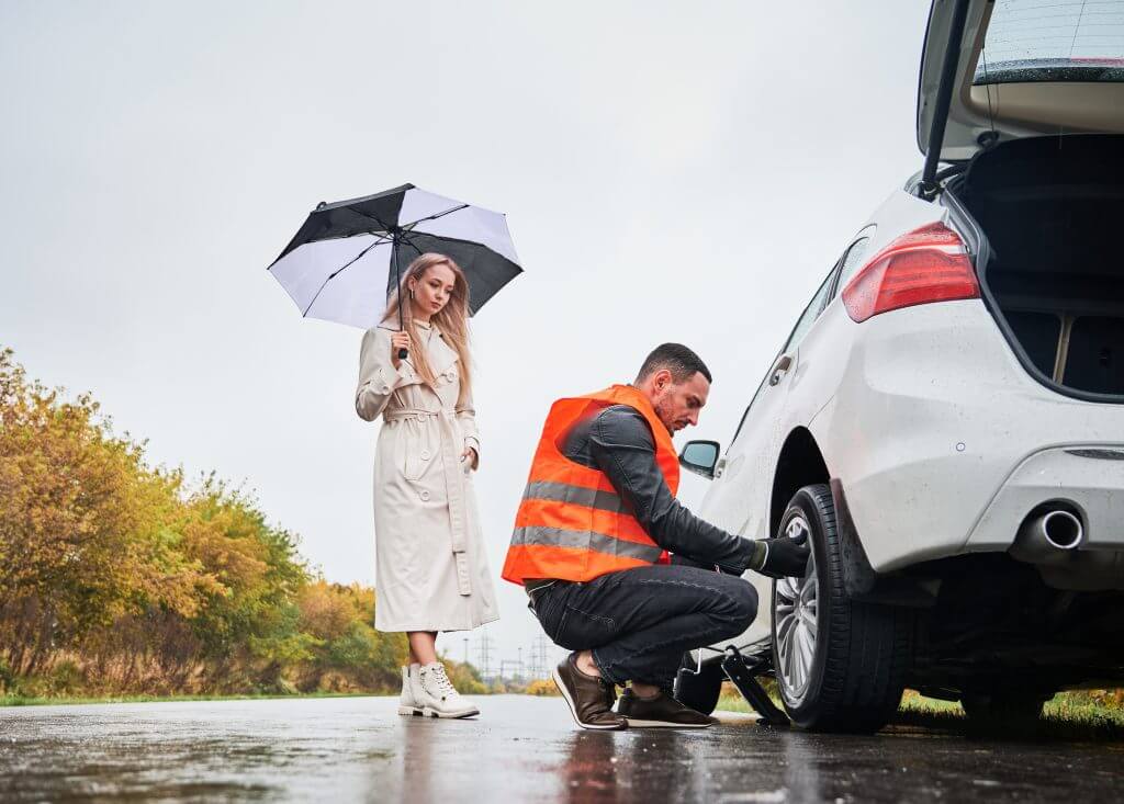 Roadside assistance worker replacing flat tire while beautiful woman in trench coat holding umbrella. Male auto mechanic changing flat tire on woman car the road. Concept of emergency road service.