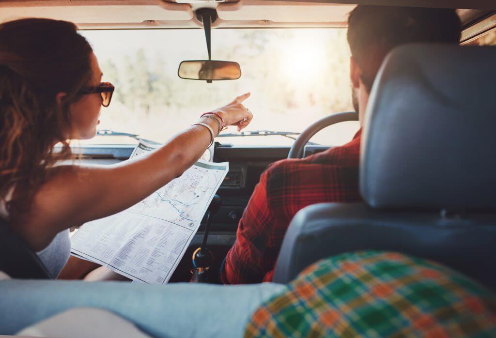 Rear view shot of couple driving on country road. Woman holding map and showing the route map to her boyfriend driving the car. Couple on a road trip.