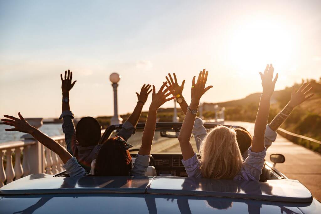 Back view of happy young friends driving cabriolet with raised hands on sunset