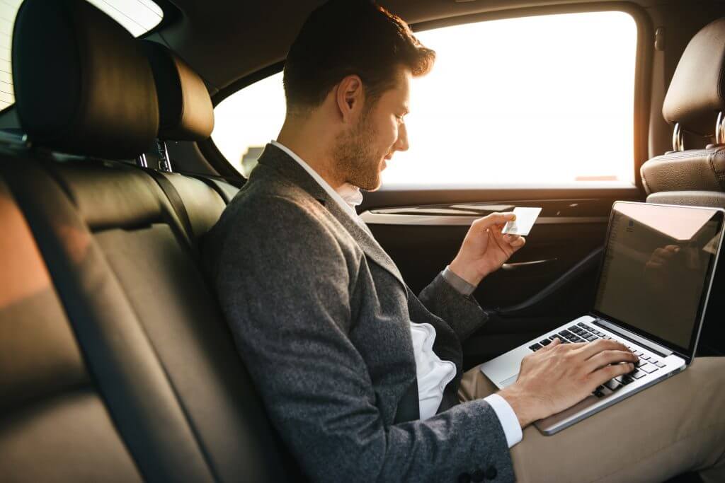 Attractive businessman using plastic credit card while sitting with laptop computer at the back of a car