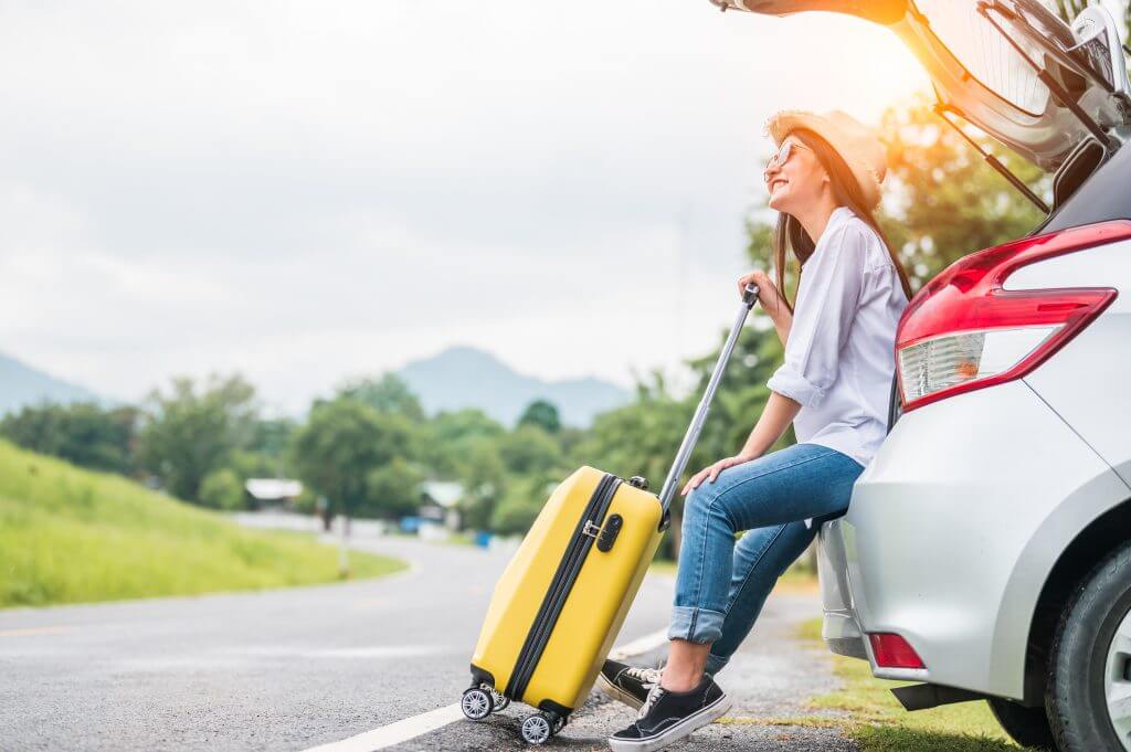 Asian woman spending weekend in roadtrip with yellow luggage. Girl relaxing on back of car with road background. Road trip and holiday vacation concept. People lifestyles and transportation.