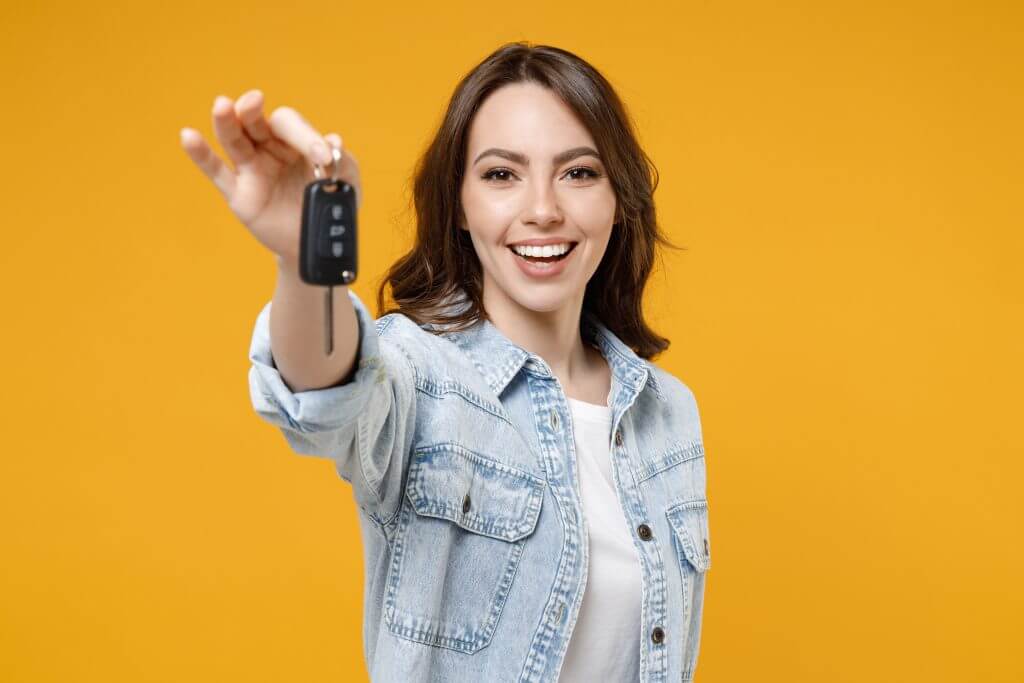 Young smiling happy satisfied excited successful brunette woman 20s in denim shirt white t-shirt hold in hands giving car keys show thumb up like gesture isolated on yellow background studio portrait