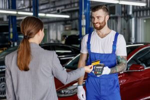 Young happy cheerful professional car mechanic man in blue overalls gloves hold payment terminal fow paying with credit card by female owner driver woman work in vehicle repair shop workshop indoors.