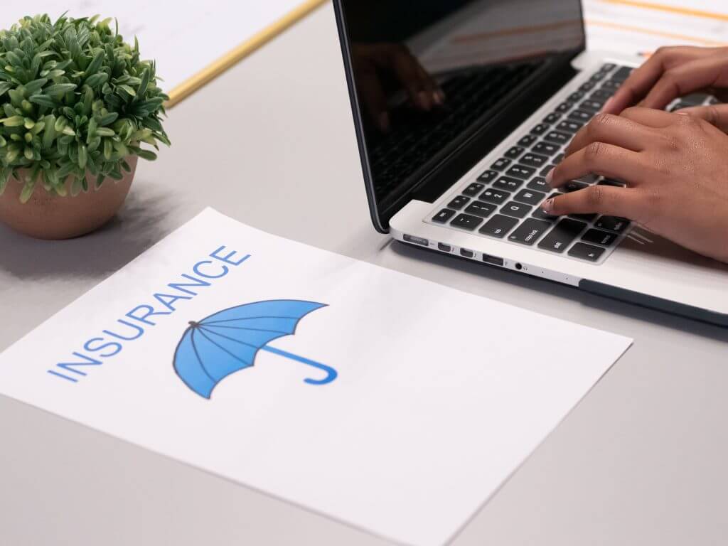 Desk photo with hands typing on a laptop and a photo of an umbrella on a bondpaper with the word "insurance" 