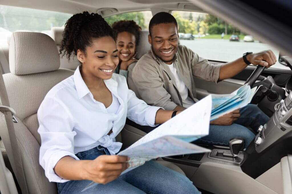 Happy Black Family Sitting In Car Looking At Road Map Planning Summer Trip. African American Parents And Daughter Choosing Destination Traveling Together. Auto Tourism Concept. Selective Focus