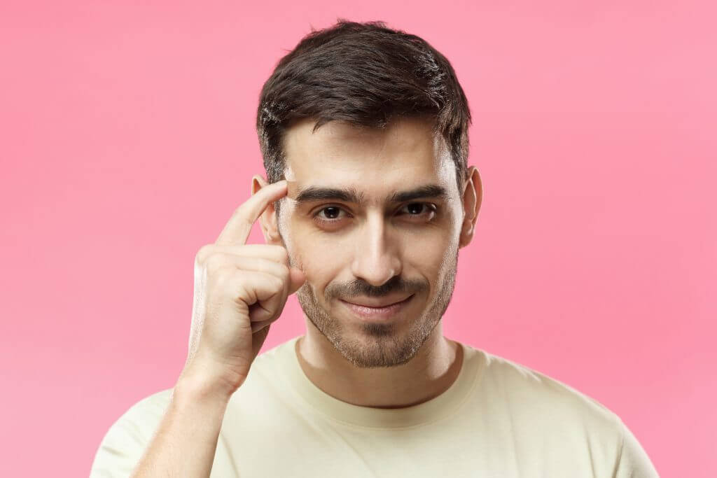 Closeup portrait of young European Caucasian man pictured isolated on pink background pressing finger to temple as if making viewer think more about offer or analyze information better for their good.
