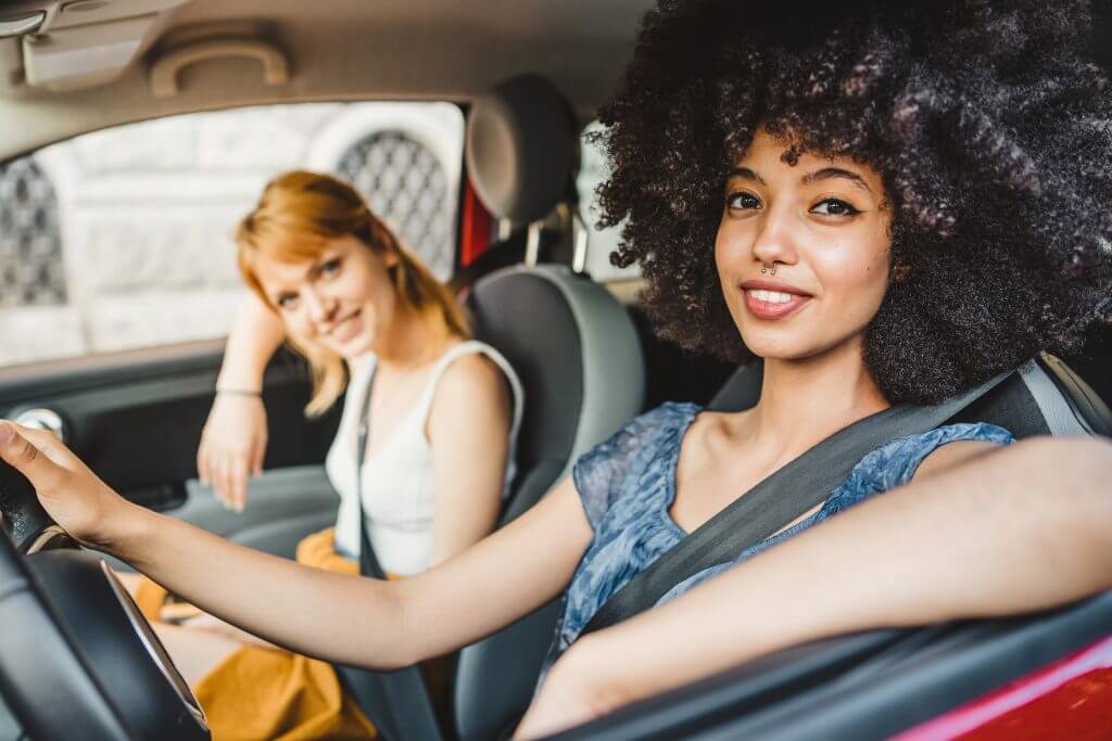 Two young women friends driving in the city - Millennials use the car to get around - People look towards the camera