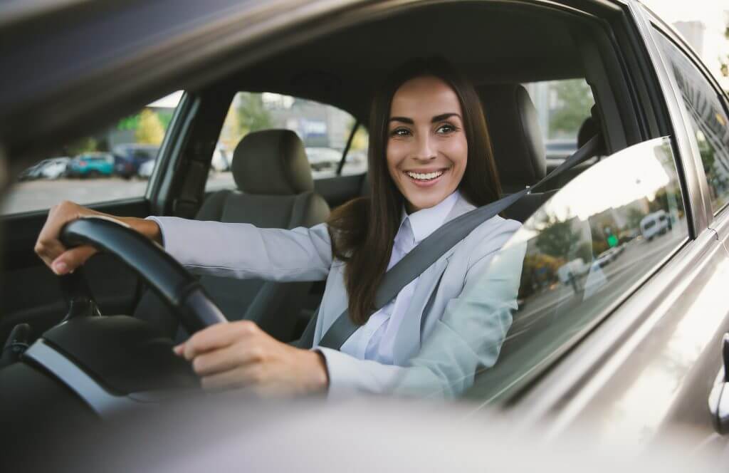 Beautiful happy successful businesswoman is driving a new modern car in good mood. Portrait cute female driver steering car with safety belt